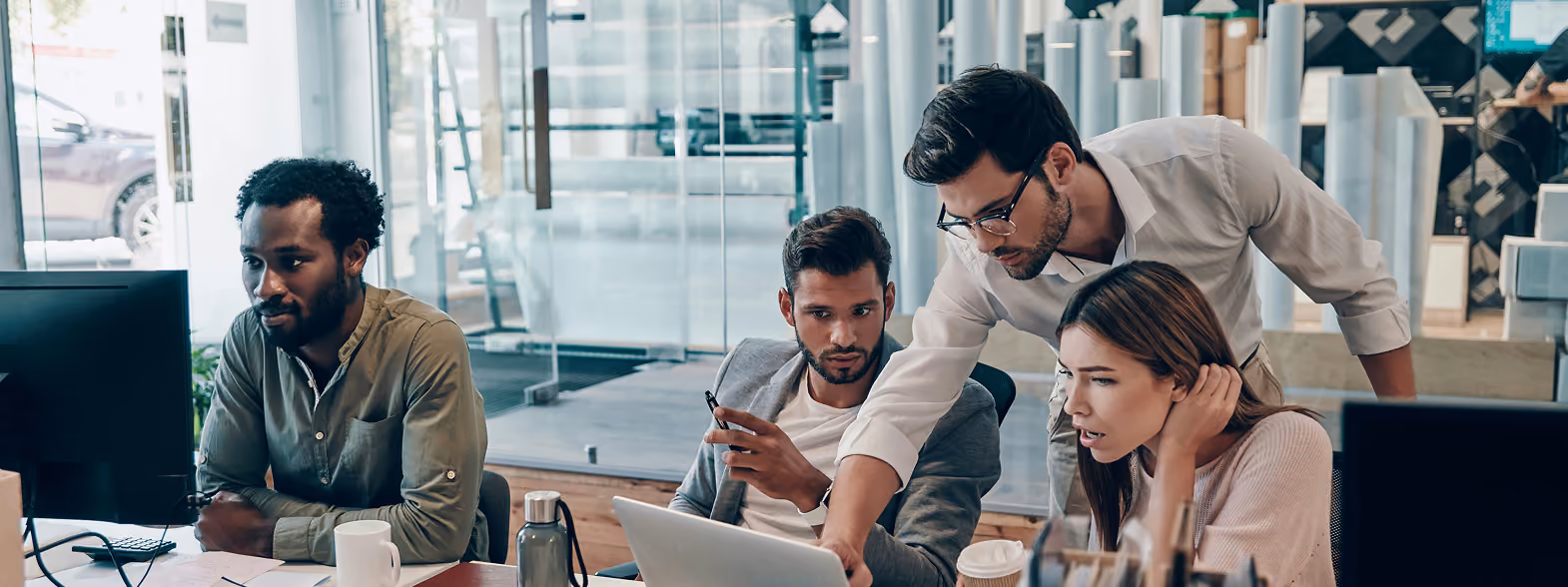 Quatre jeunes professionnels concentrés travaillent ensemble devant des ordinateurs dans un bureau moderne, discutant et examinant un écran d'ordinateur portable.