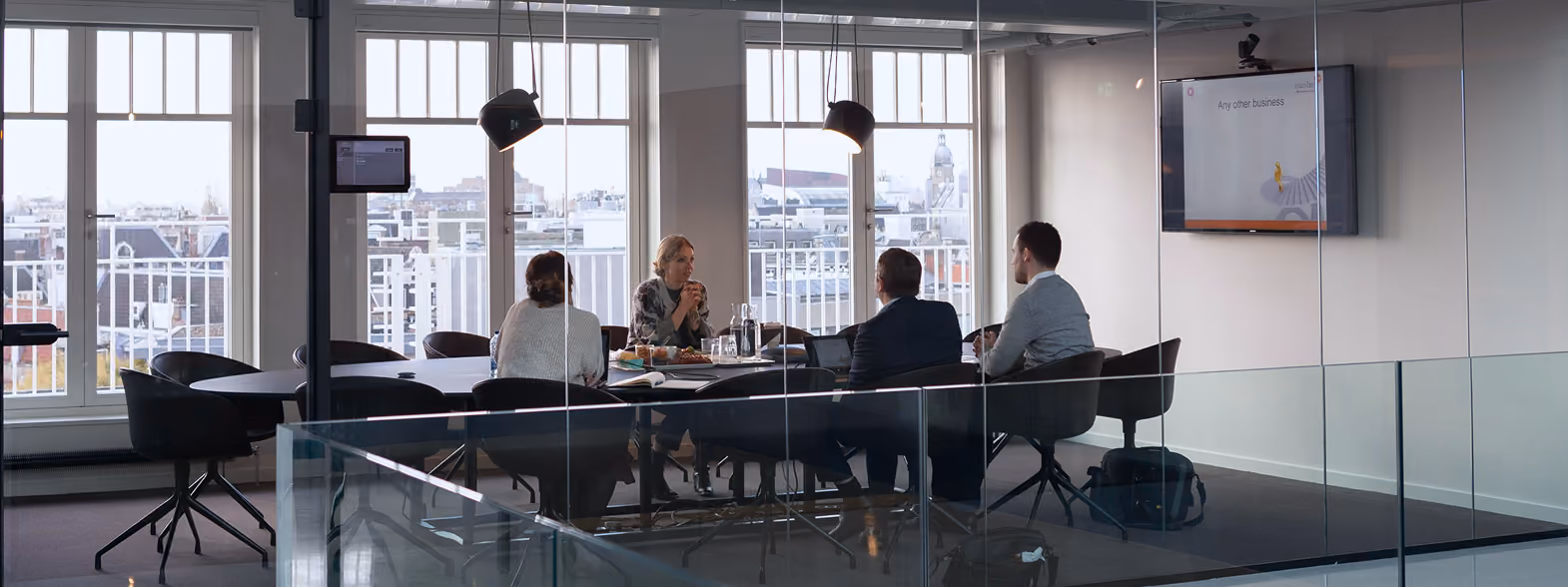 Quatre personnes en réunion autour d'une table dans une salle de conférence lumineuse avec grandes fenêtres donnant sur un paysage urbain, un écran de présentation fixé au mur, et plusieurs chaises noires.