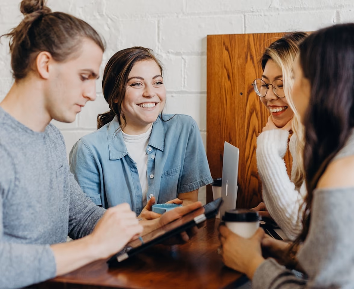 Quatre jeunes adultes assis autour d'une table en bois dans un café, discutant et souriant, avec un ordinateur portable et des boissons chaudes.
