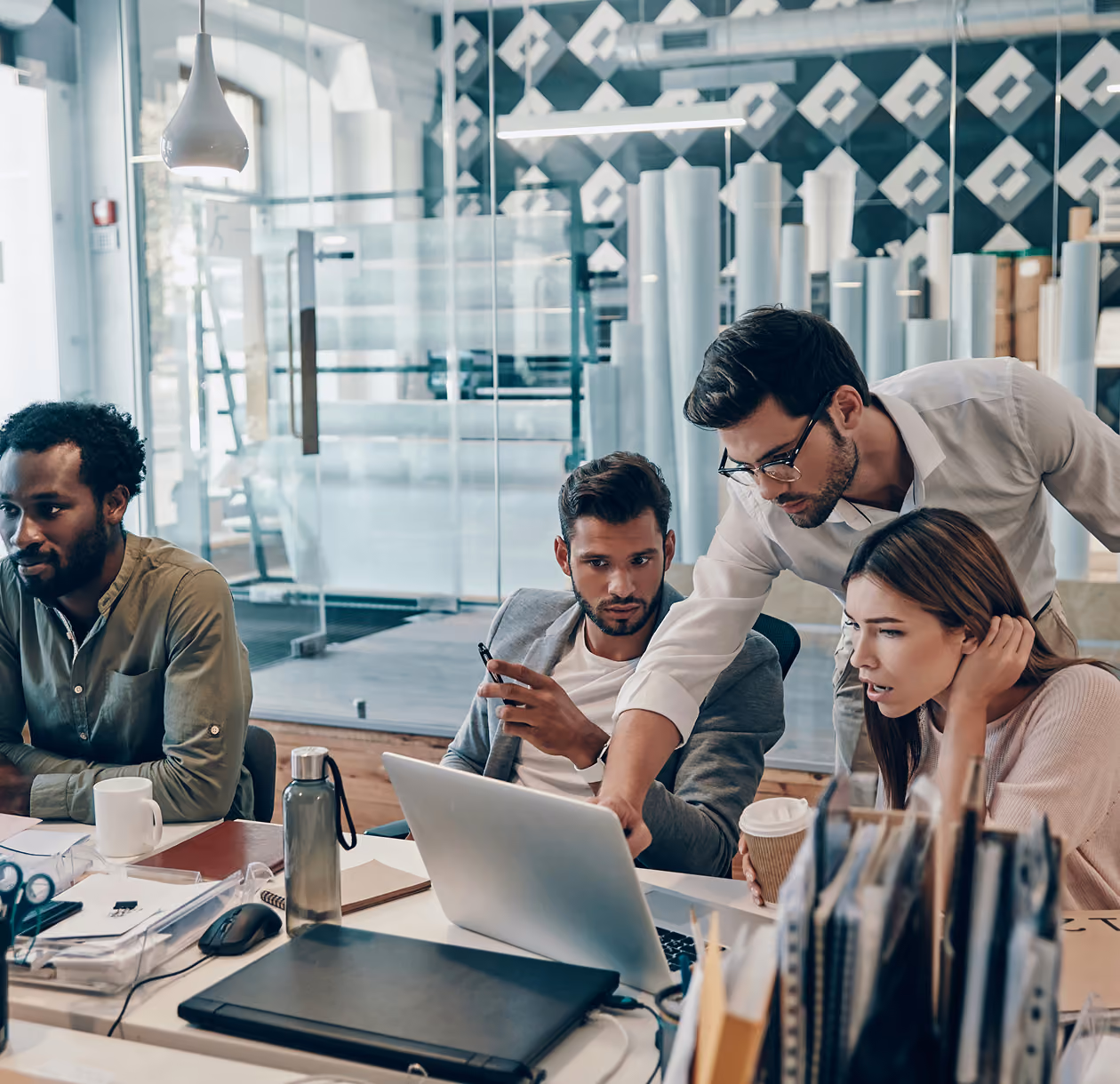 Quatre personnes concentrées collaborent devant un ordinateur portable dans un bureau moderne avec des dossiers, tasses et bouteilles sur la table.