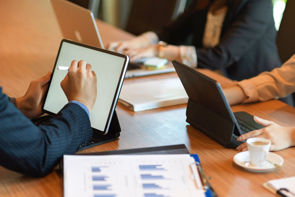 Personne en costume utilisant un stylet sur une tablette posée sur une table en bois, avec deux autres personnes travaillant sur des ordinateurs portables et tablettes autour de la table, et une tasse de café à côté.
