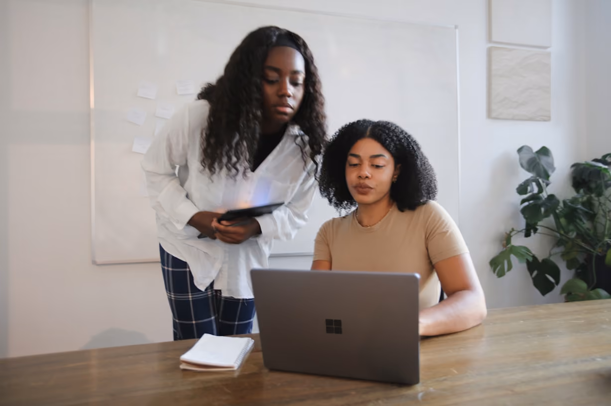 Deux femmes travaillant ensemble devant un ordinateur portable dans une salle de réunion avec un tableau blanc en arrière-plan.