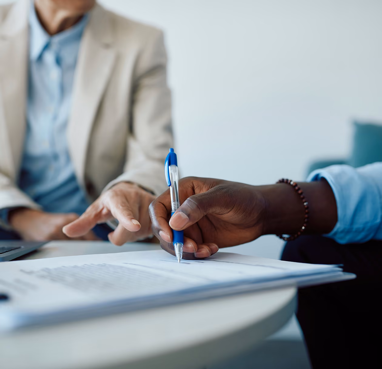 Une main tenant un stylo bleu au-dessus d'un document sur une table, tandis qu'une autre personne en arrière-plan pointe vers le document.