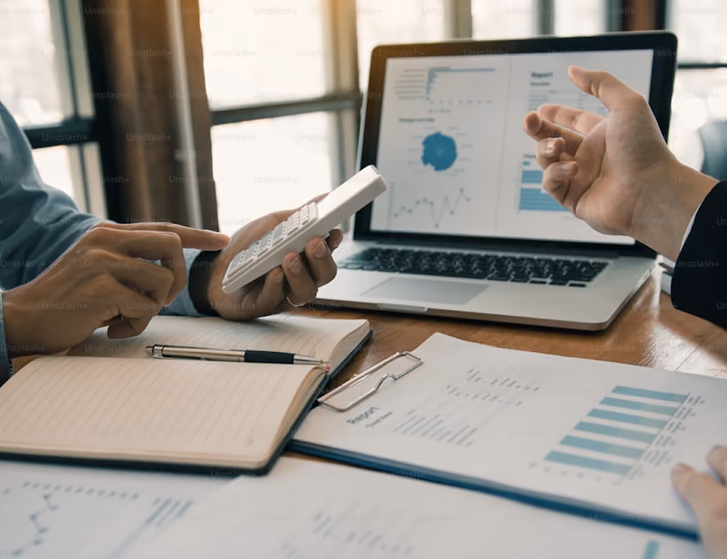 Deux personnes travaillant ensemble avec un ordinateur portable montrant des graphiques et des rapports, un carnet ouvert avec un stylo, une calculatrice et des documents imprimés avec des graphiques sur une table en bois.