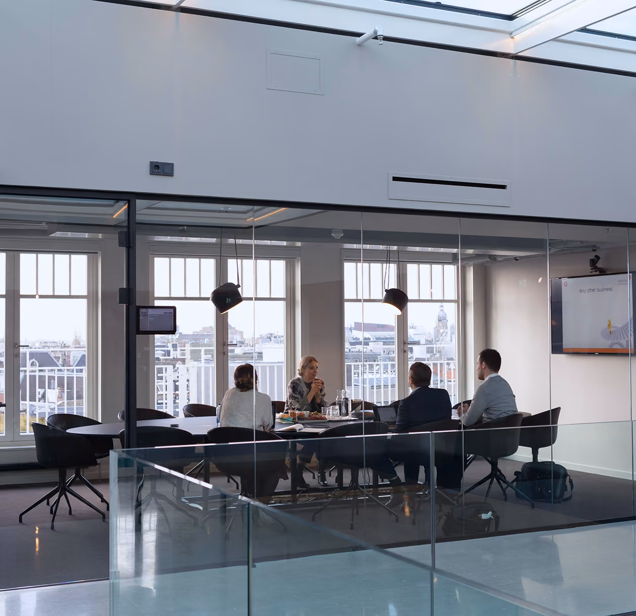 Quatre personnes assises autour d'une table dans une salle de réunion vitrée avec vue sur une ville depuis de grandes fenêtres, éclairage suspendu et un écran sur le mur affichant une présentation.