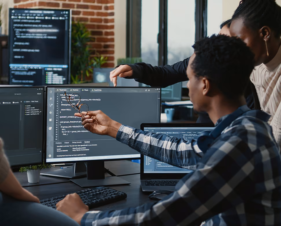 Un groupe de personnes travaille ensemble dans un bureau moderne, examinant du code informatique affiché sur plusieurs écrans d'ordinateur, l'un d'eux tenant des lunettes devant un écran principal avec du code visible.