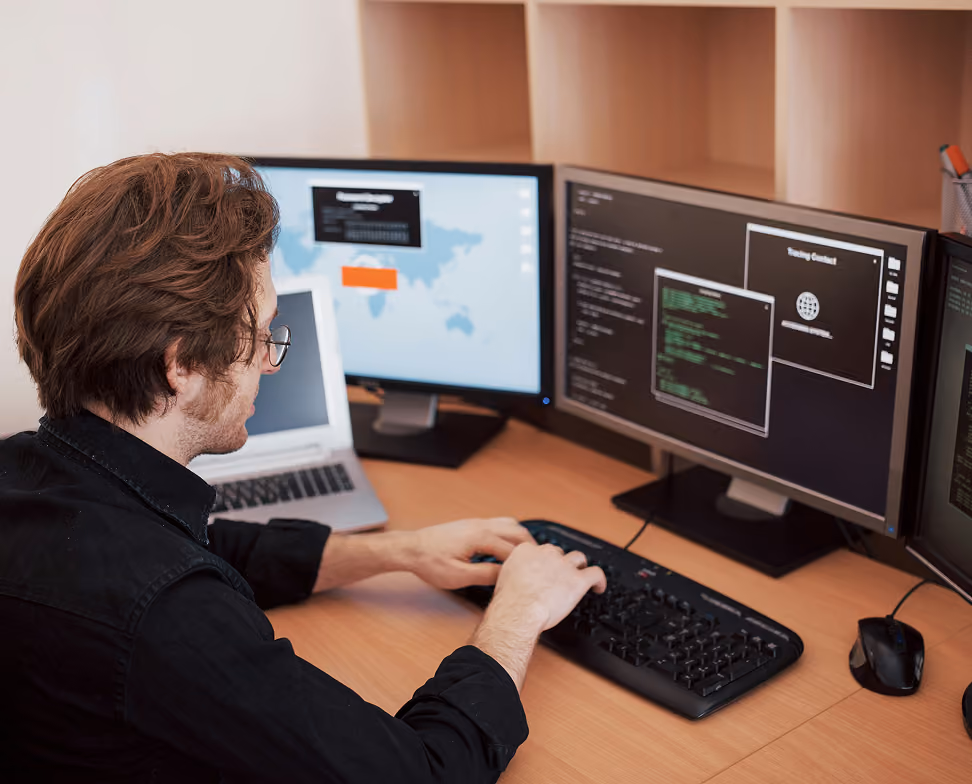 Homme avec des lunettes assis à un bureau, tapant sur un clavier d’ordinateur, face à deux écrans affichant du code informatique et une carte du monde, avec un ordinateur portable fermé à côté.