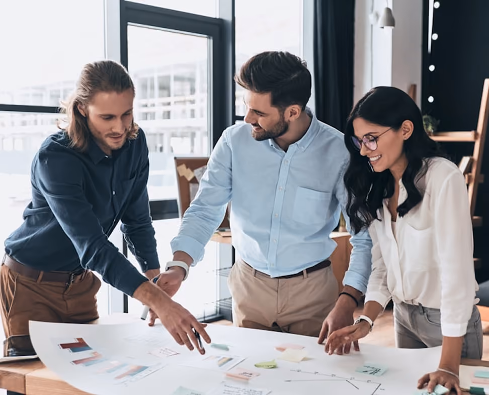 Trois jeunes professionnels collaborent autour d'une grande table où sont étalés des documents et des graphiques, discutant et pointant les informations affichées.