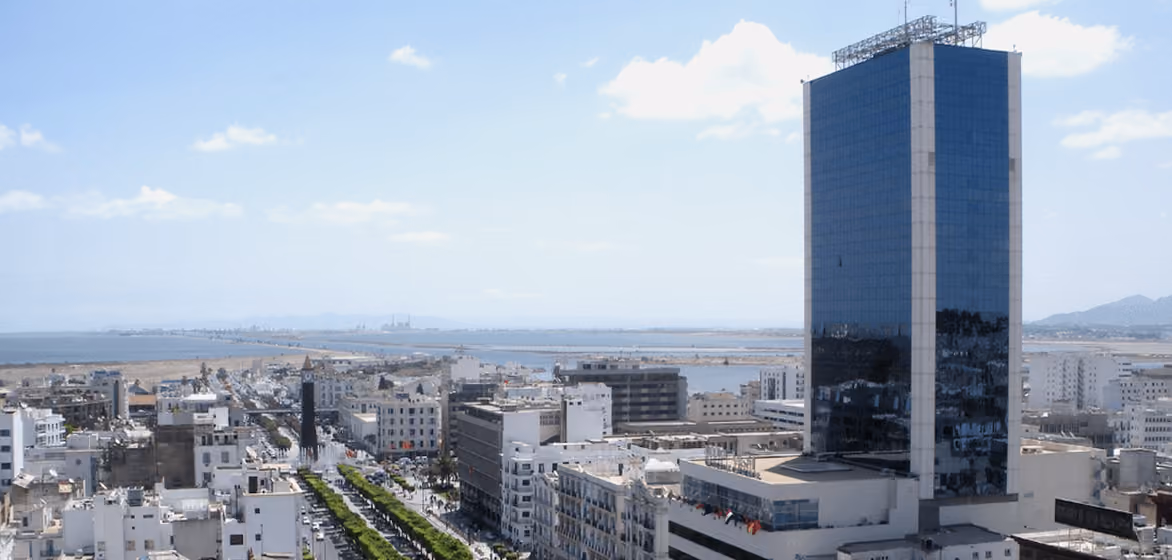 Vue panoramique d'une partie de la ville de Tunis avec des bâtiments modernes et anciens, une grande tour en verre à droite, une avenue arborée au centre, et la mer en arrière-plan sous un ciel bleu avec quelques nuages.