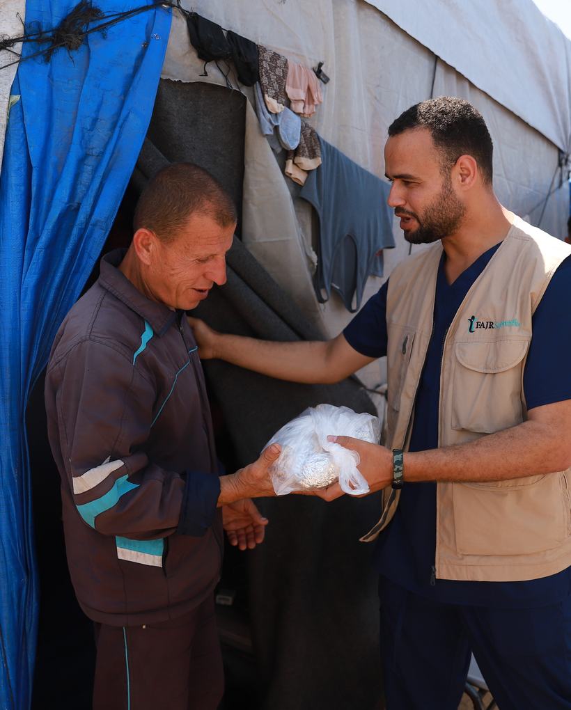 A FAJR aid worker handing supplies to a man outside a displacement shelter.