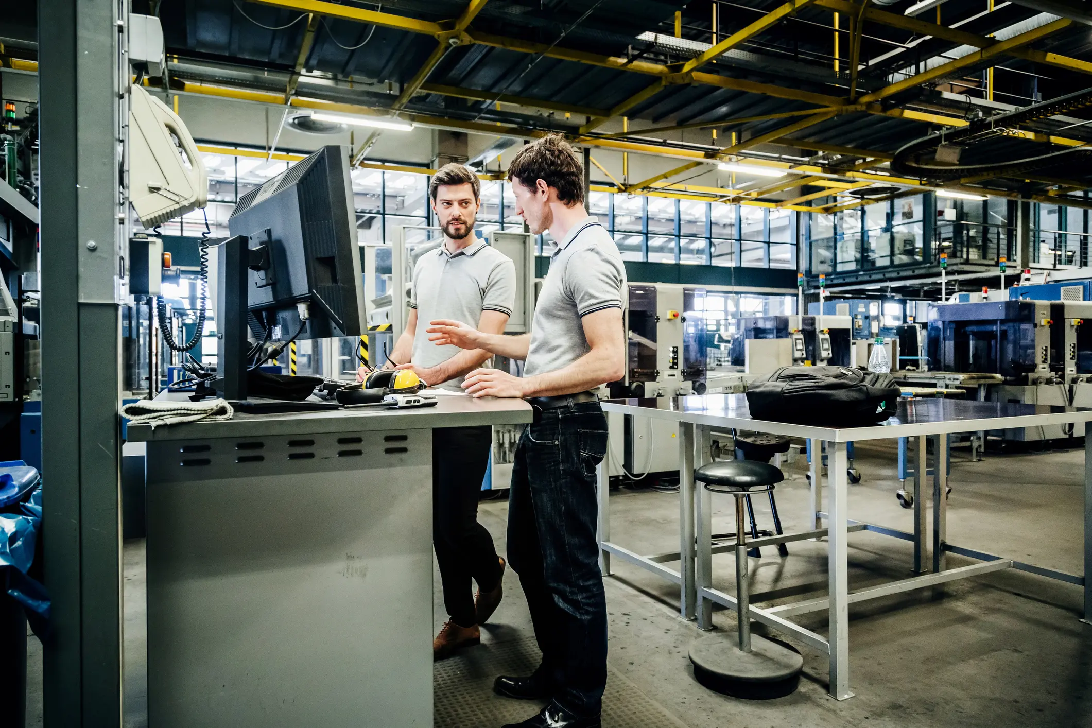 Twee mannen in polo's overleggen bij een computerscherm in een fabriekshal met industriële machines en werkbanken.