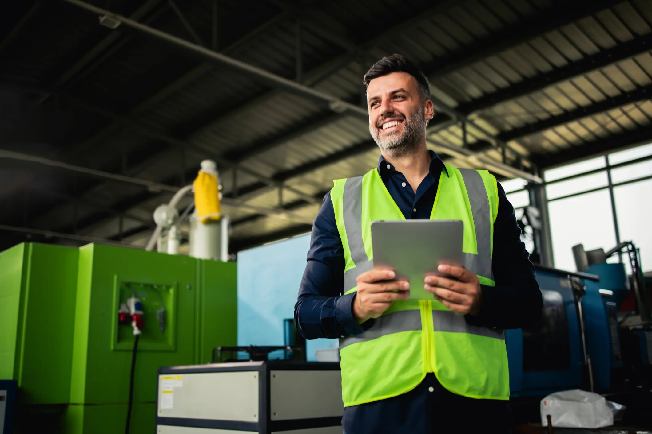 Tevreden man met een fluorescerend geel veiligheidsvest, staand in een fabriekshal, die een tablet vasthoudt.