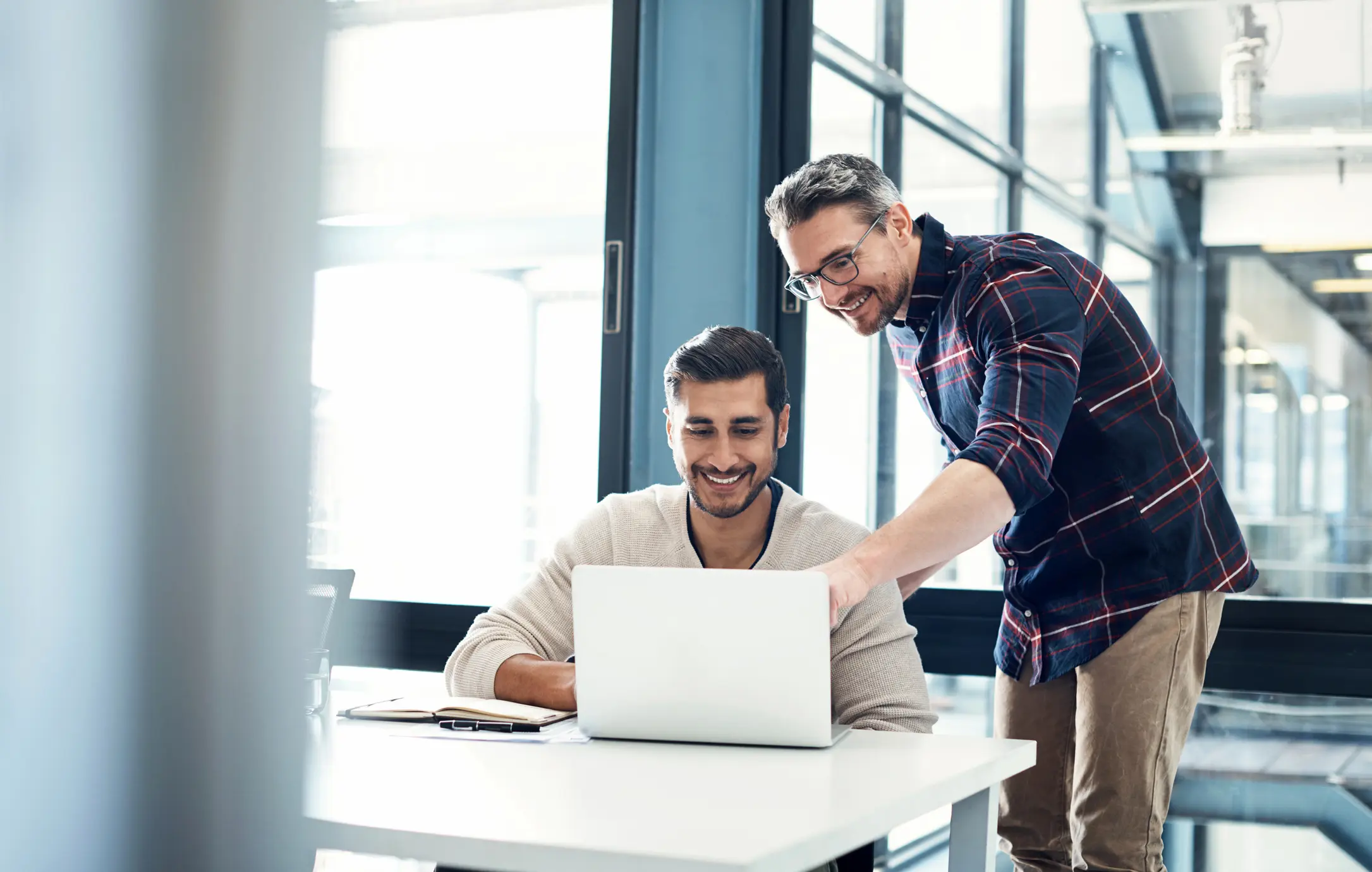 Twee mannen werken samen in een modern kantoor, waarbij de staande man iets op de laptop van de zittende man aanwijst terwijl ze beiden glimlachen.