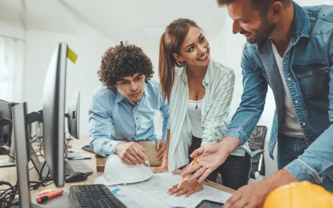 Drie jonge professionals bespreken bouwplannen aan een bureau met bouwtekeningen, een computer en bouwhelmen.