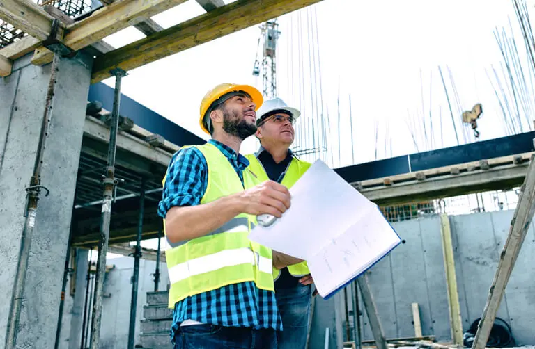 Twee bouwvakkers met veiligheidsvesten en helmen bekijken bouwplannen op een bouwterrein met betonstructuren en metalen staven.