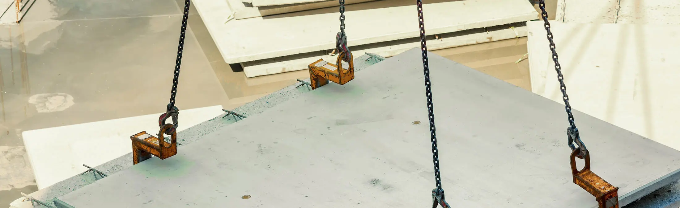 Betonnen plaat wordt hijsbaar ondersteund door drie metalen kettingen met oranje hijshaken op een bouwplaats met andere betonplaten op de achtergrond.