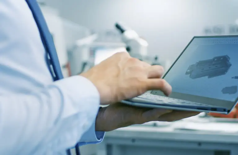 Een persoon in een laboratoriumjas werkt op een laptop met een 3D-model van een technisch onderdeel op het scherm.