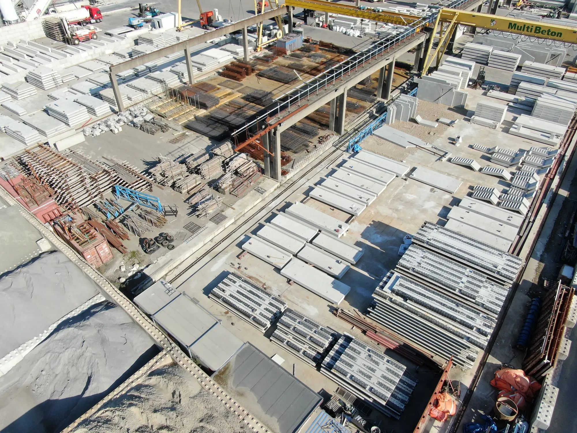 Luchtfoto van een bouwplaats met grote stapels prefab betonnen panelen, metalen staven en bouwmaterialen onder een beginfase constructie met een geel hijskraan met opschrift 'Multi Beton'.