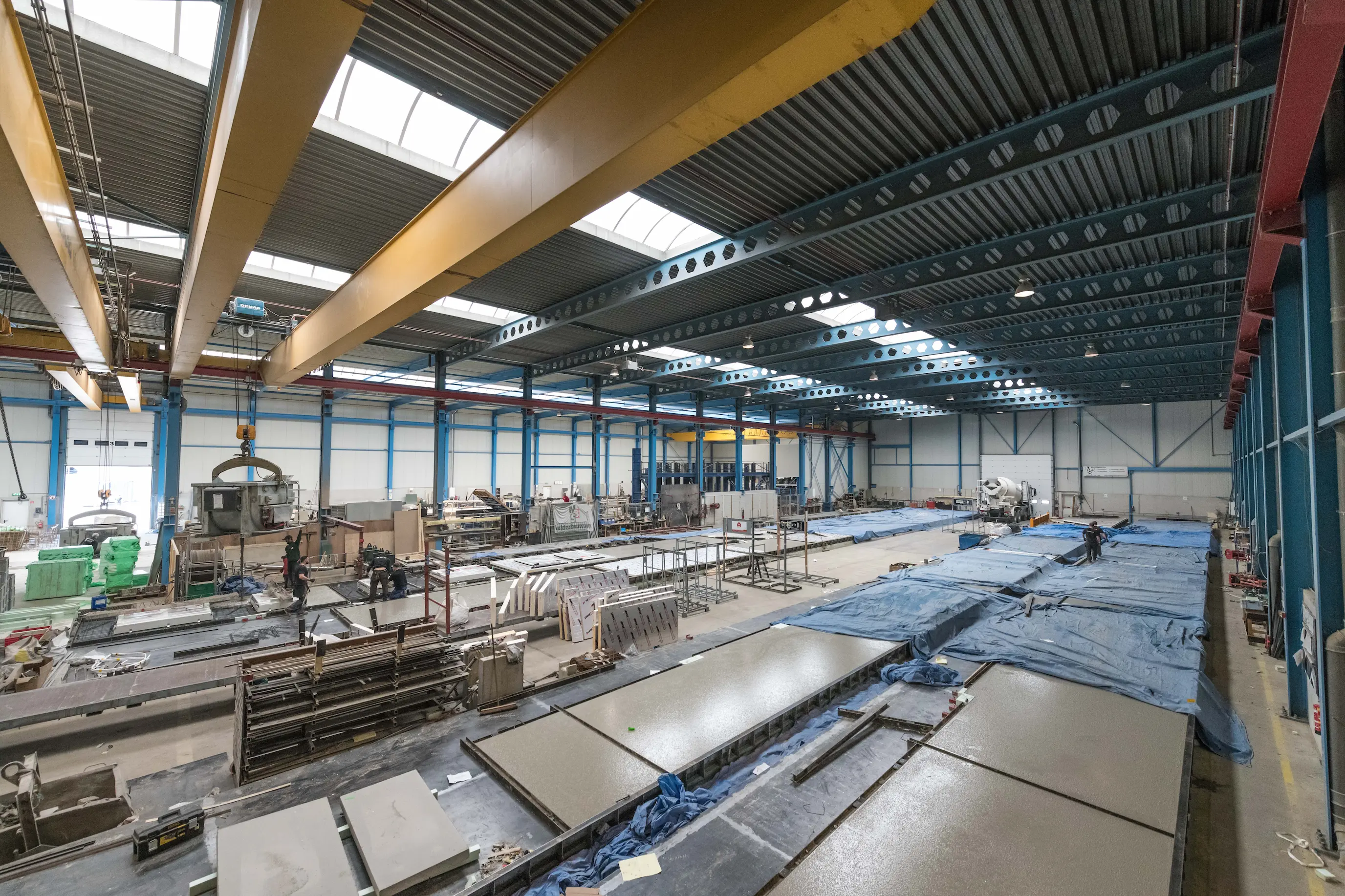 Grote fabriekshal met afgewerkte betonnen platen en arbeiders die betonproducten maken onder een metalen dak met grote gele hijsarmen.