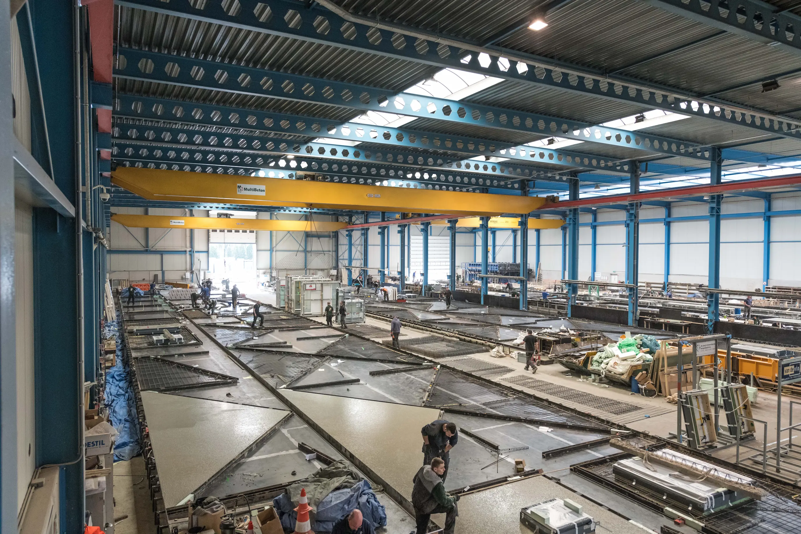 Grote fabrieksruimte waar meerdere arbeiders bezig zijn met het gieten en afwerken van grote betonnen platen in metalen vormen, met gele hijskranen en blauwe staalconstructies aan het plafond.