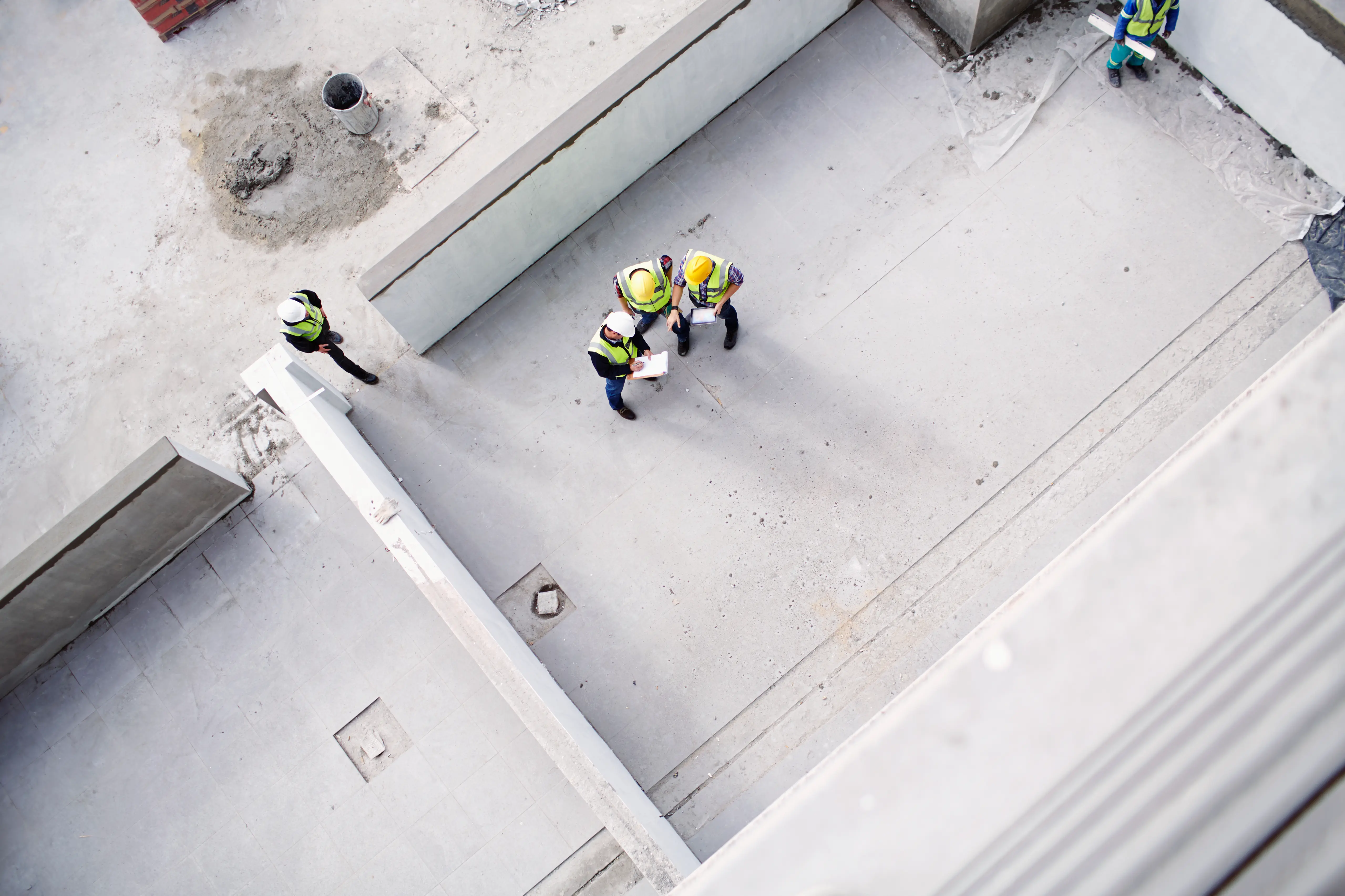 Vier bouwvakkers met helmen en reflecterende vesten staan op een bouwplaats met betonnen muren en vloeren, drie bespreken documenten en een staat apart.