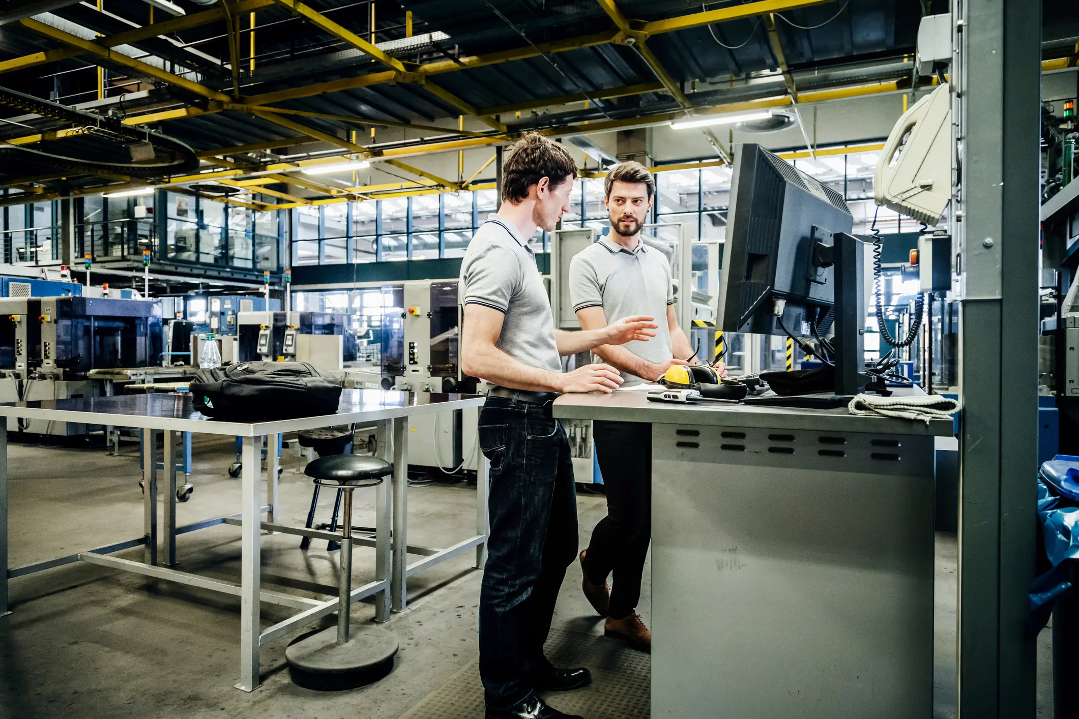 Twee mannen in een industrieel magazijn of fabriek bespreken iets terwijl ze bij een computer staan met machines op de achtergrond.