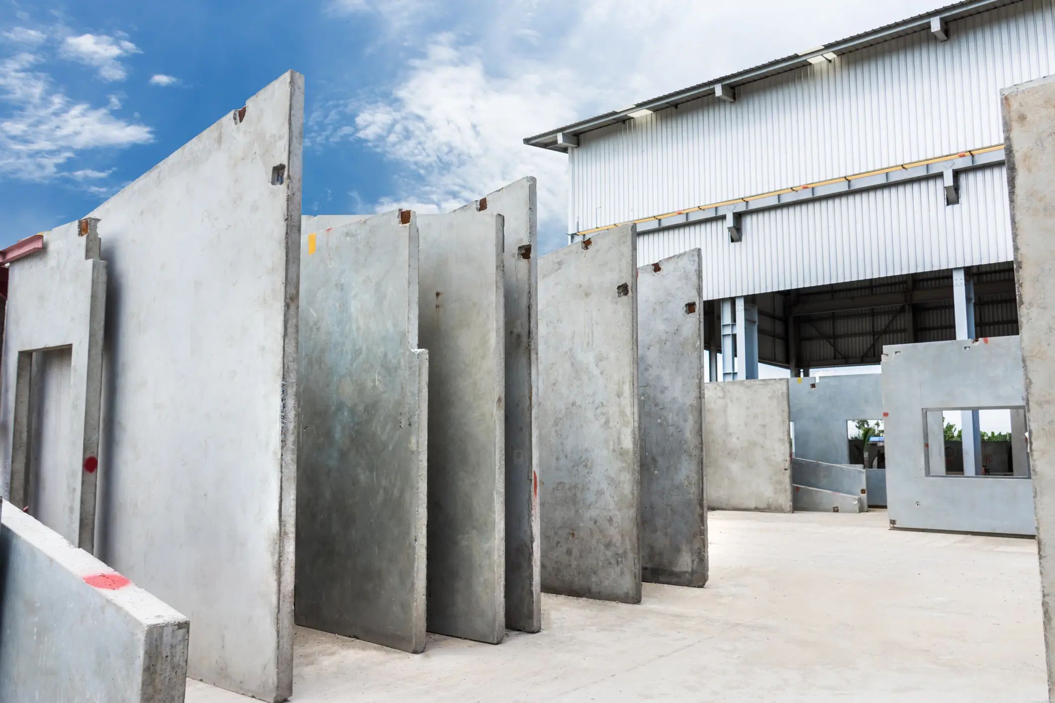 Vooraf vervaardigde betonnen wanden staan verticaal opgesteld in een fabriekshal onder een blauwe lucht met enkele wolken.