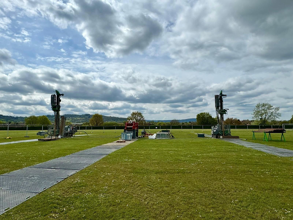 Two borehole drilling rigs on a grassy field with ground protection mats.