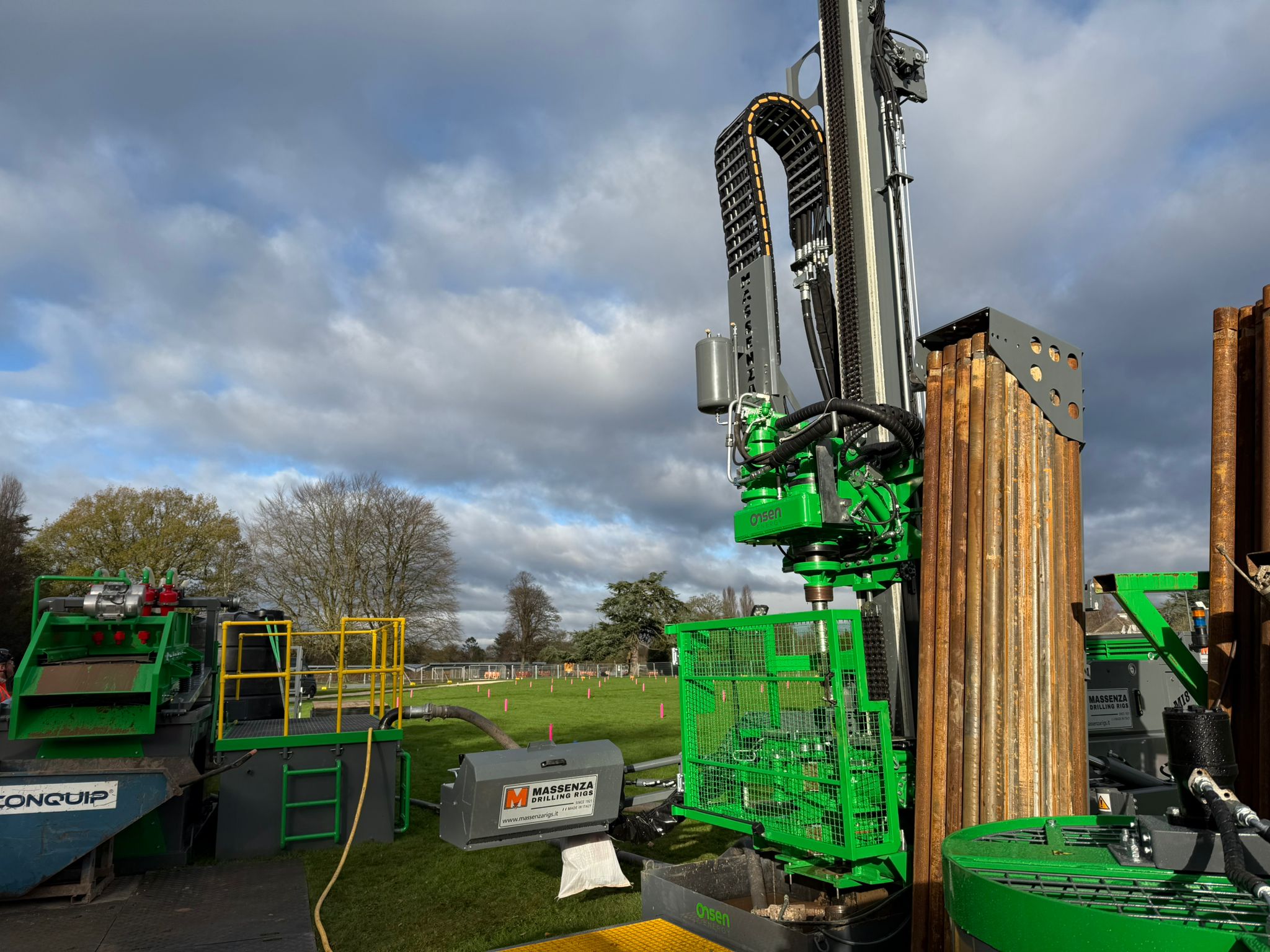 Green Massenza drilling rig on a grassy site with a rack of borehole casings.