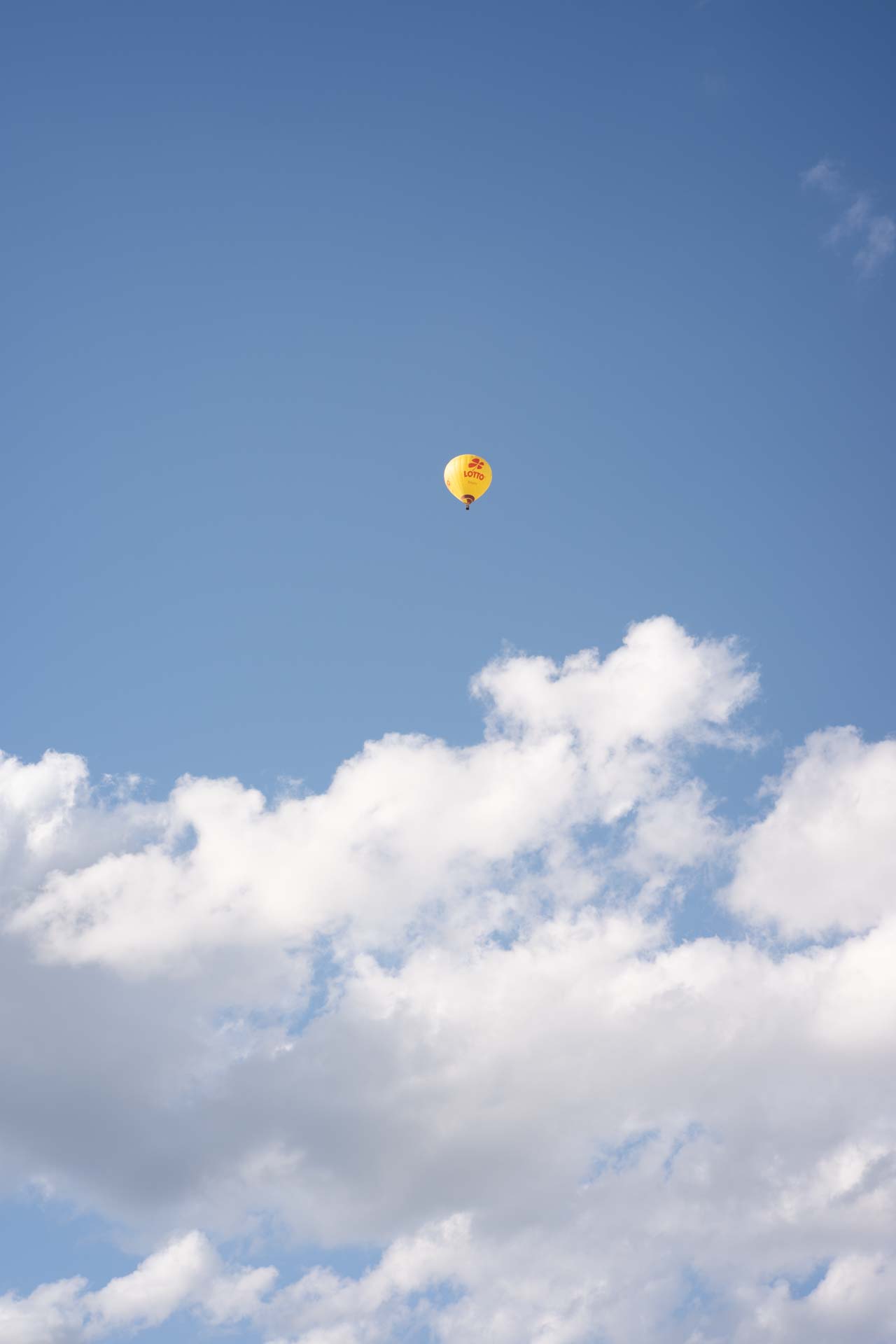 Yellow hot air balloon with red markings floating high in a blue sky above white clouds.