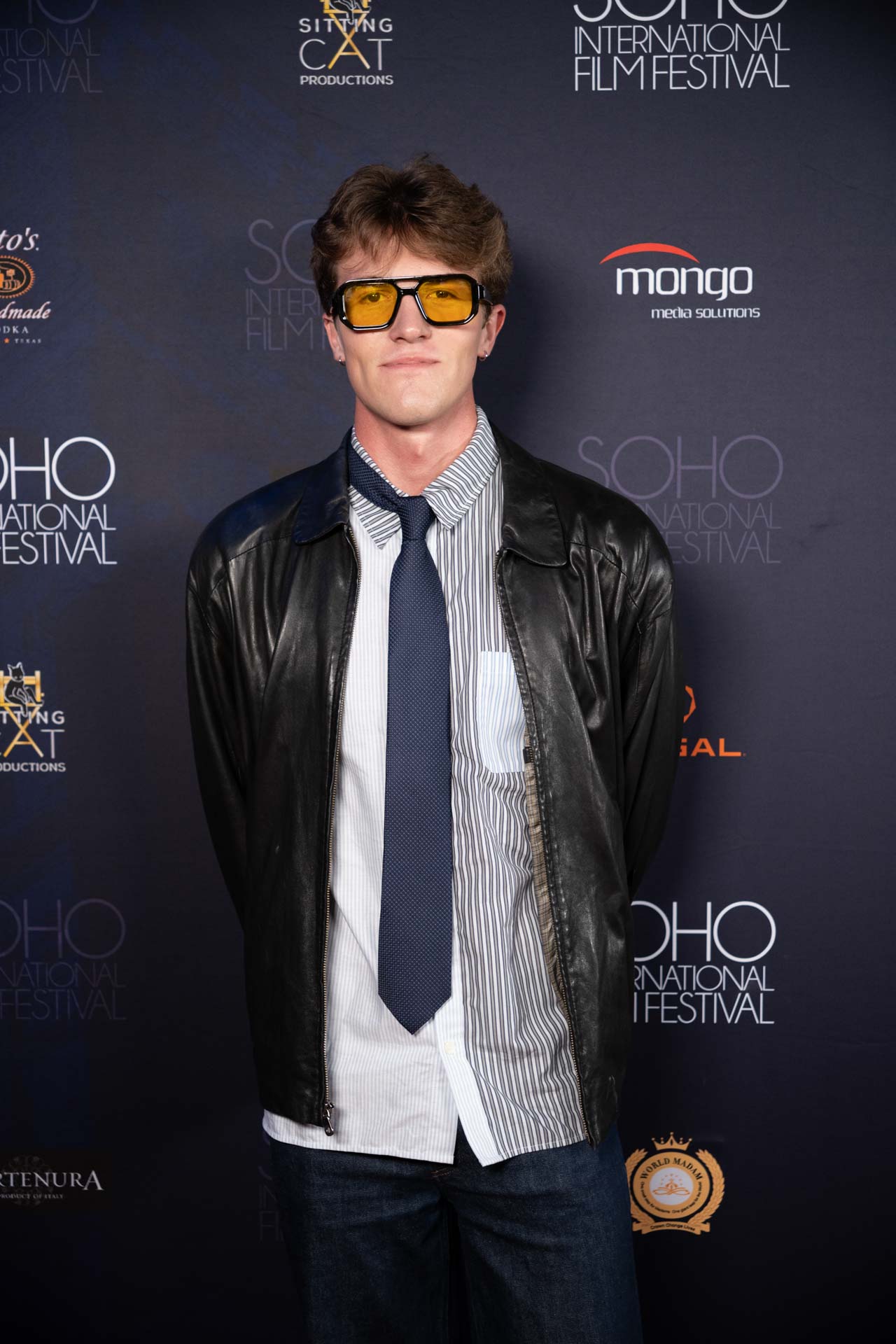 Young man wearing yellow-tinted sunglasses, a striped shirt with a navy tie, and a black leather jacket standing against a Soho International Film Festival backdrop.