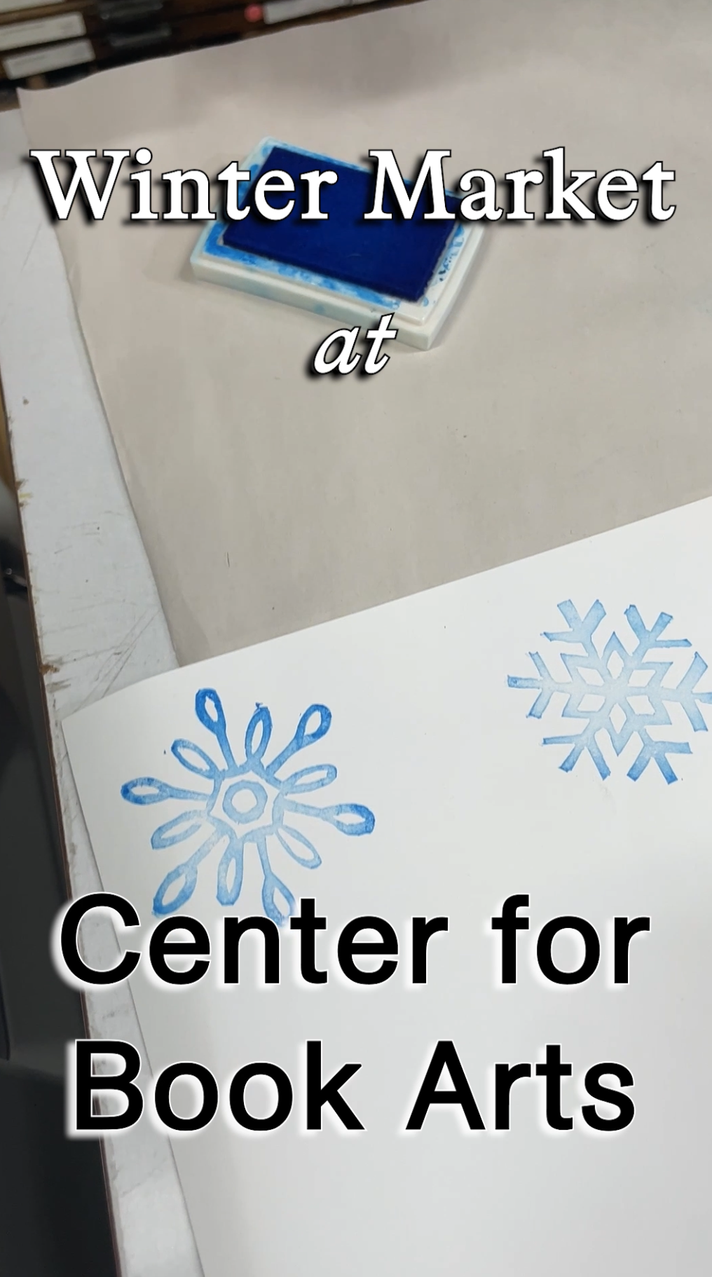 A blue ink stamp pad on a white table next to a sheet of paper with two blue stamped snowflake designs, overlaid with text reading 'Winter Market at Center for Book Arts'.
