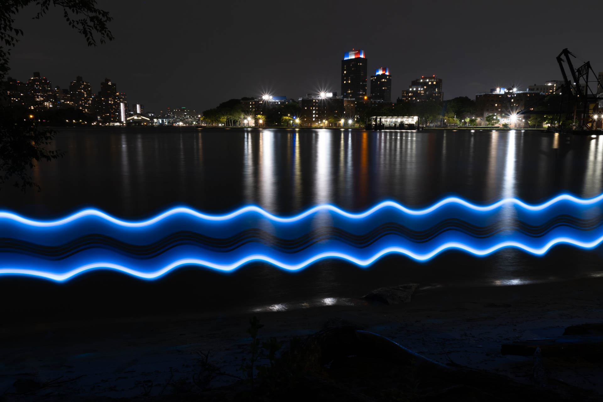 Night cityscape across water with blue wavy light trails reflecting on the surface.