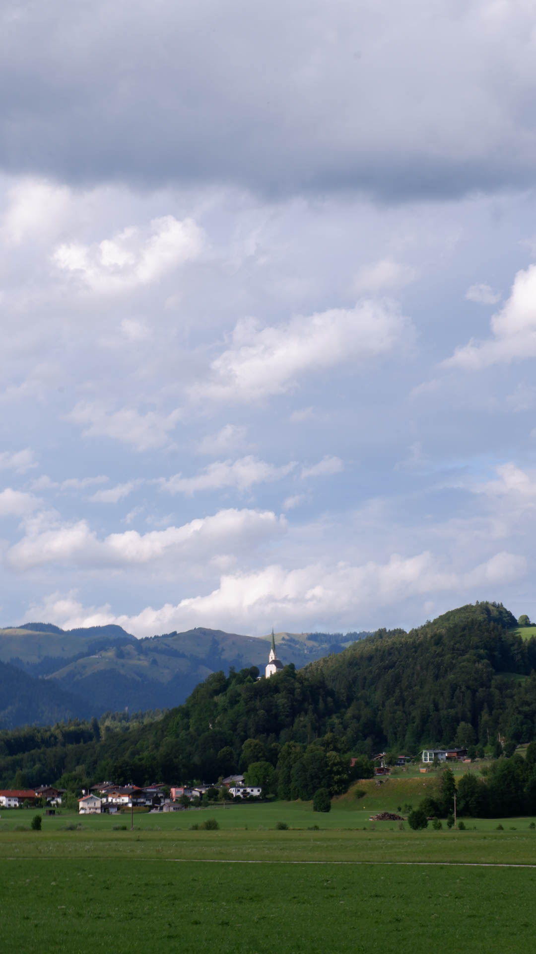 Green fields and forested hills with a small white church on a hill under a cloudy blue sky.