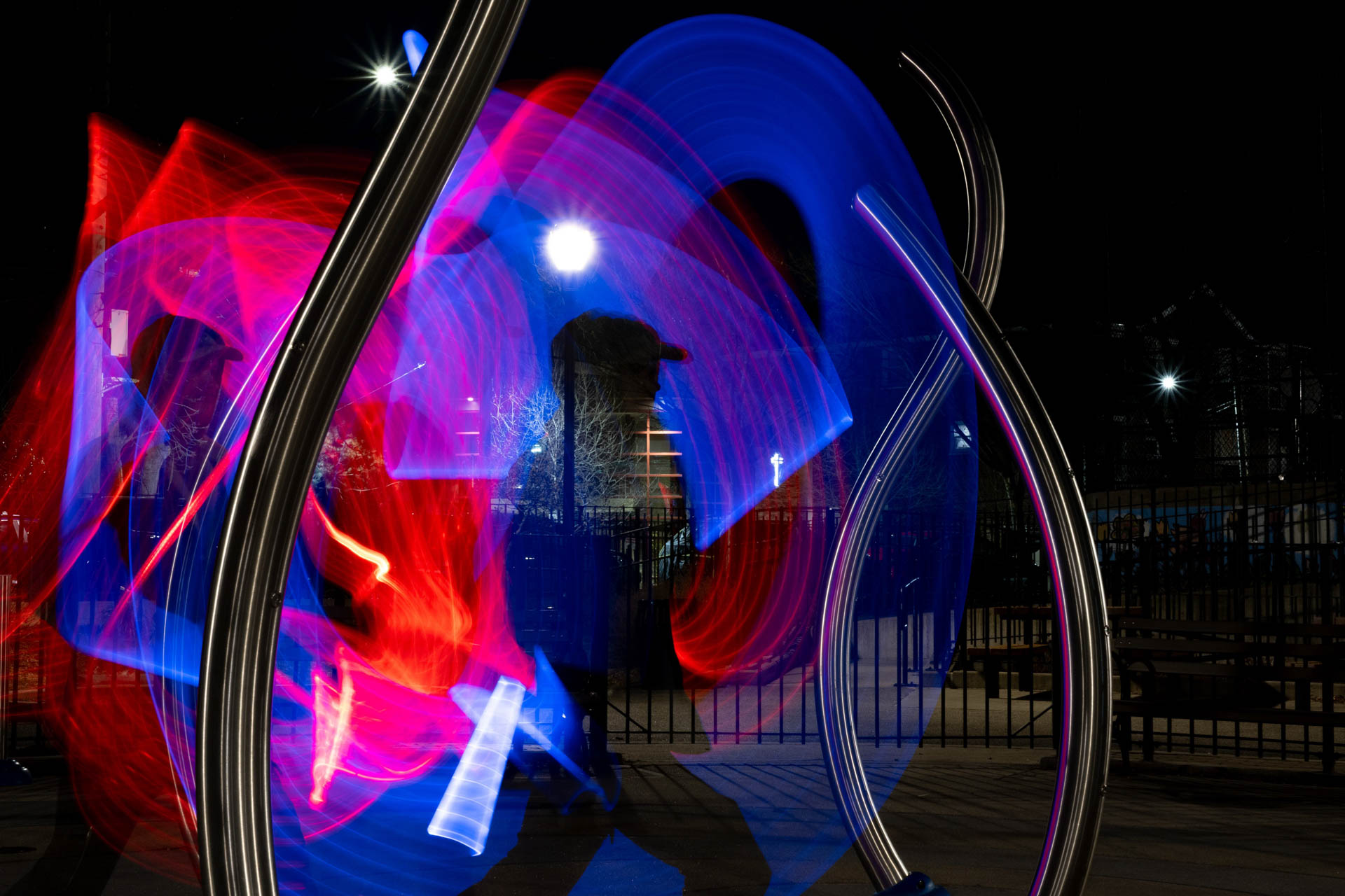 A person in a dark outdoor setting surrounded by swirling red and blue light trails and two large curved silver metal structures at night.