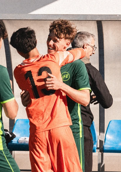 Two soccer players hugging in celebration, one wearing an orange jersey with the number 12 and the other in a green jersey with yellow trim, standing near blue plastic seats and a person in black clothing in the background.
