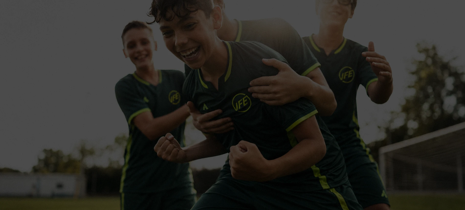 Group of four young soccer players in dark green uniforms with yellow trim celebrating on a field near a goalpost, one player in front is clenching fists and smiling.