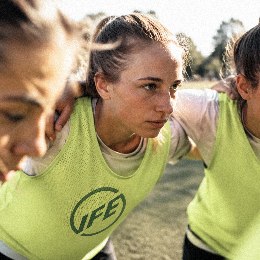 Three young women in yellow sports vests huddle closely outdoors, preparing or strategizing for a game or activity, with focused expressions.