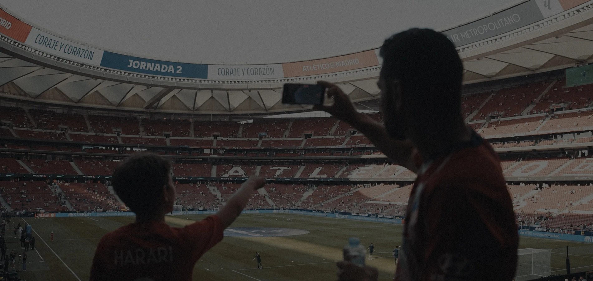 Two soccer fans in red jerseys inside a large stadium, one pointing towards the field and the other taking a photo with a phone, with rows of empty and occupied seats in the background.