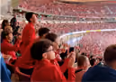 Fans in a stadium, mostly wearing red, cheering and clapping while watching an event.