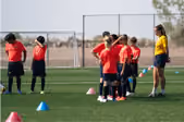 A youth soccer team in red jerseys and black shorts gathers on a green field with scattered cones, while a woman in a yellow shirt and black shorts stands nearby, possibly coaching.