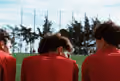 Three people wearing red outfits and orange neck warmers sitting side by side outdoors with blurry trees in the background.