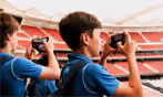 Two young boys in blue sports jerseys in a stadium, each holding and looking through a pair of binoculars.