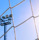 A tall sports stadium floodlight tower seen through a close-up of a net against a clear blue sky.