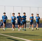 Group of six young male soccer players jogging together on a grass field during daylight, wearing blue sportswear.