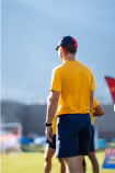 Person in a yellow shirt and dark shorts standing outdoors, wearing a red cap, facing away from the camera.