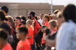 Group of children and adults outdoors, many wearing red shirts, with one child raising a hand and others gathered closely.