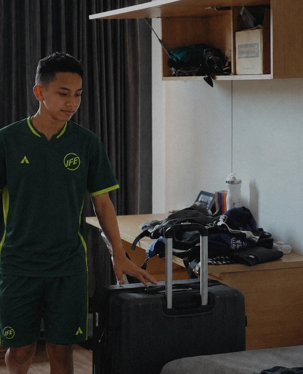 Young man in green sportswear standing indoors next to a large black suitcase with an extended handle, in a room with wooden furniture and folded clothes on a desk.