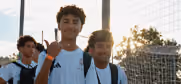Two young people outdoors with bright sunlight behind them, one making a hand gesture while smiling.