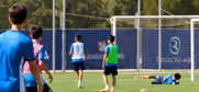 Four young male soccer players running or standing on a grass field near a goal, with one player lying on the ground and a fenced area in the background.